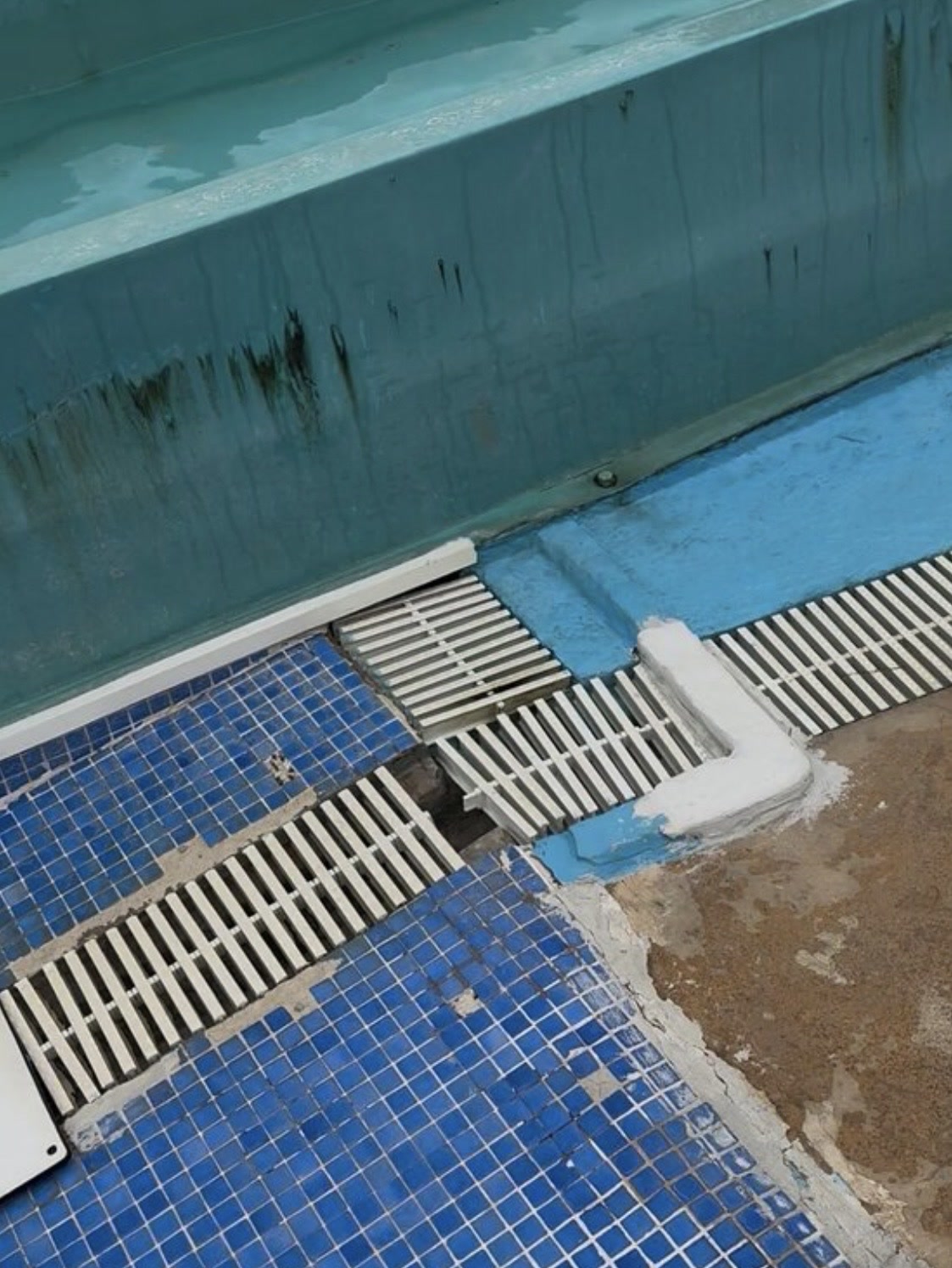 Vora de la piscina fotografiada amb peces del mosaic absents, trams de reixeta desplaçats i trencats, buits al desguàs, revestiment blau desgastat i marques fosques a la paret.